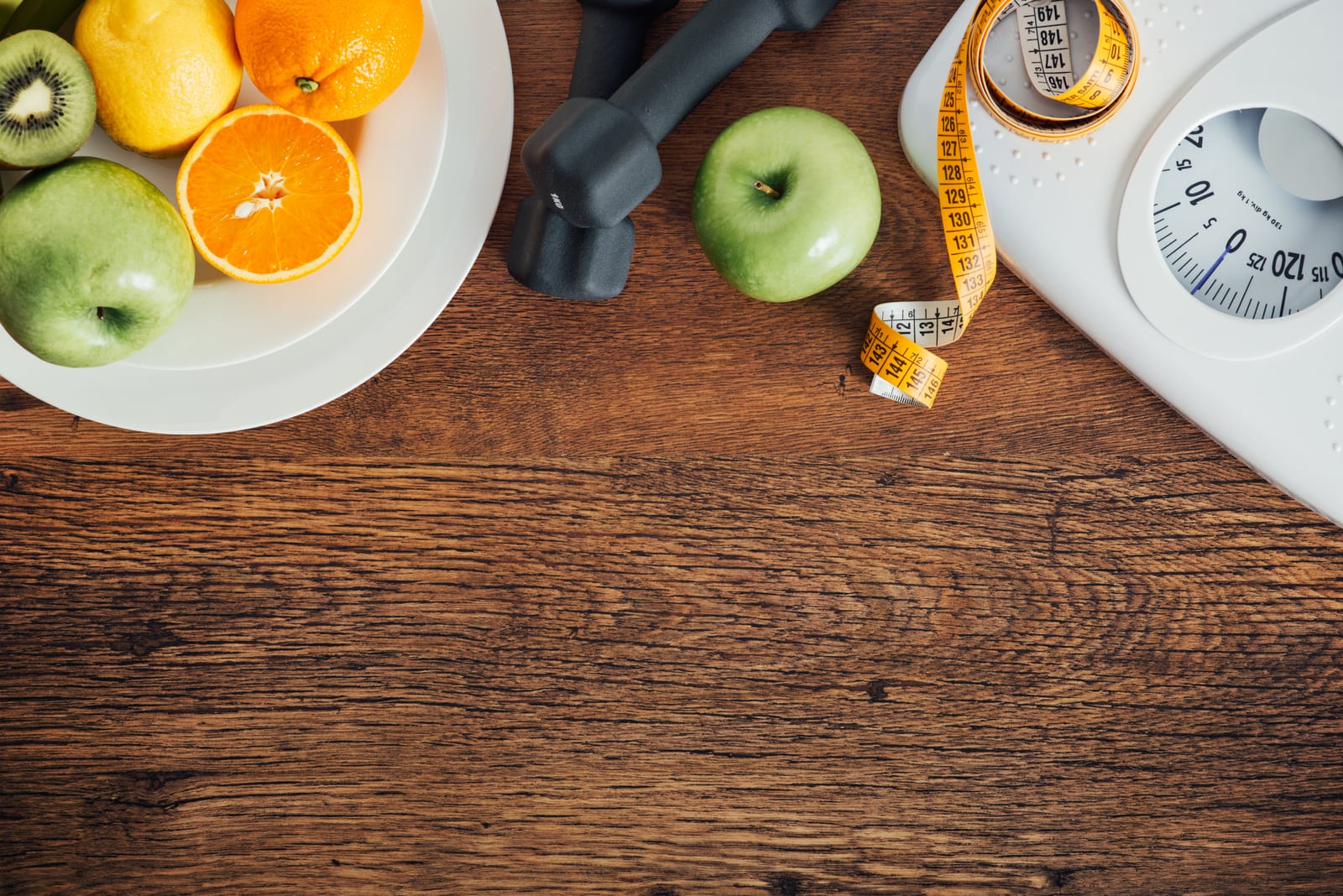 Fresh fruit, a dumbbell, a tape measure, and a bathroom scale arranged on a wooden table